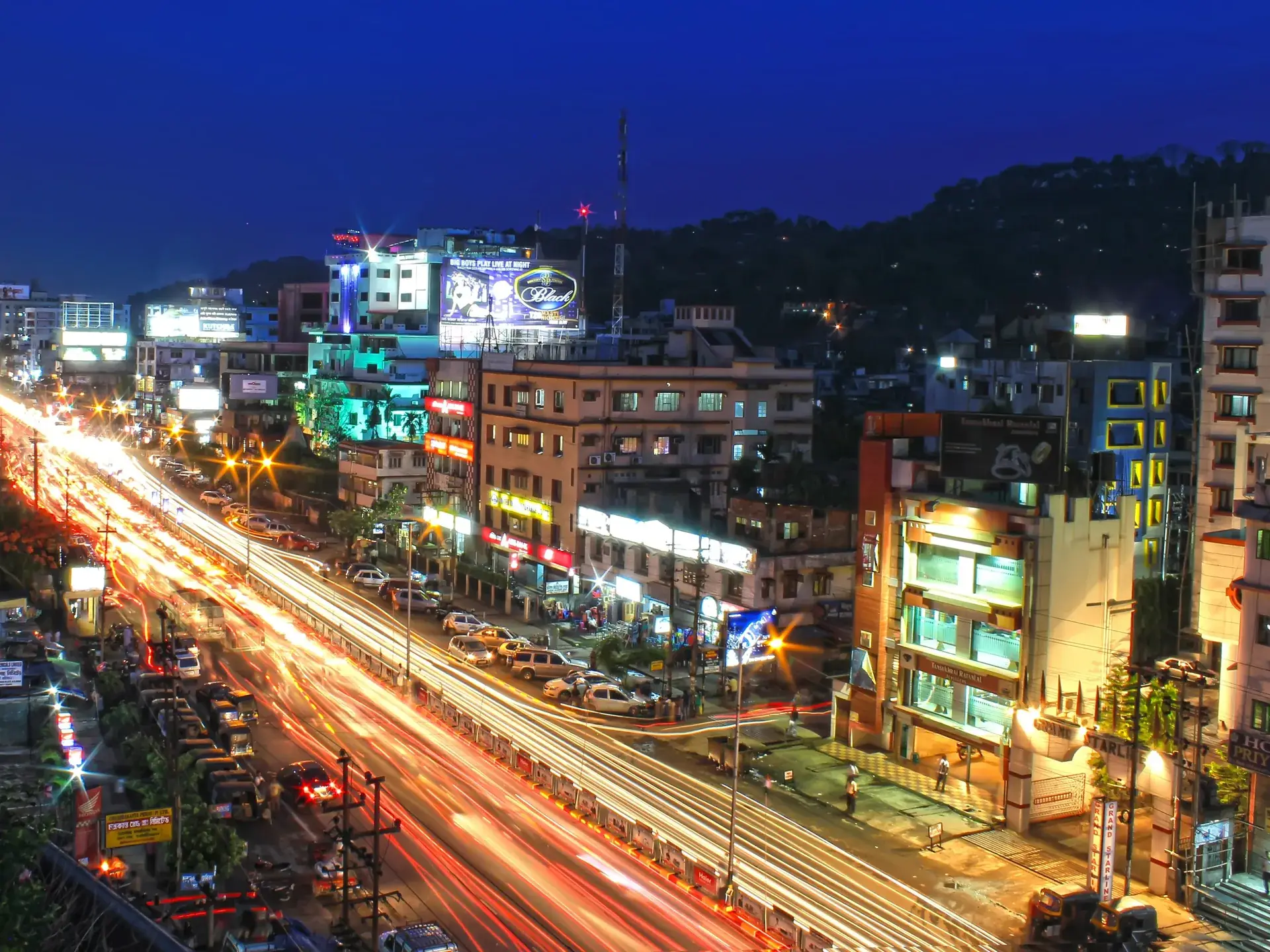 Guwahati street at dusk with light trails from traffic, lit shops and hotels, and dark wooded hills behind